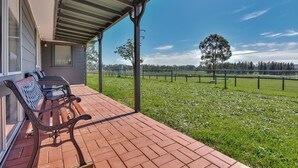 A covered outdoor area with a brick floor, a bench, and a view of a grassy field with trees. - Andres Farm Cottage (Broke)
