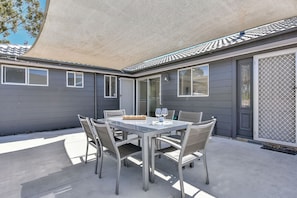 Une terrasse extérieure avec une table et des chaises, un espace abrité et une maison à l’extérieur grise.