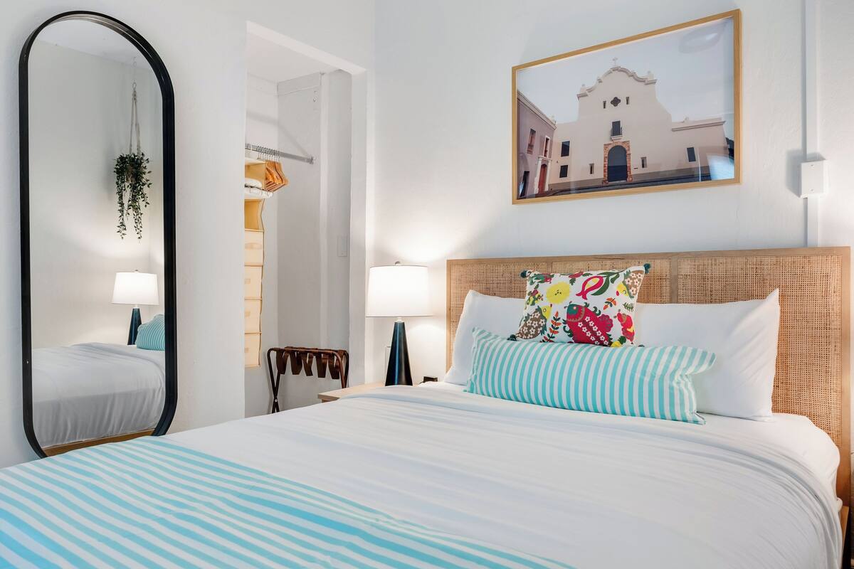 Une chambre d’hôtel avec un grand lit, un miroir, un tableau encadré représentant un bâtiment et une plante suspendue.