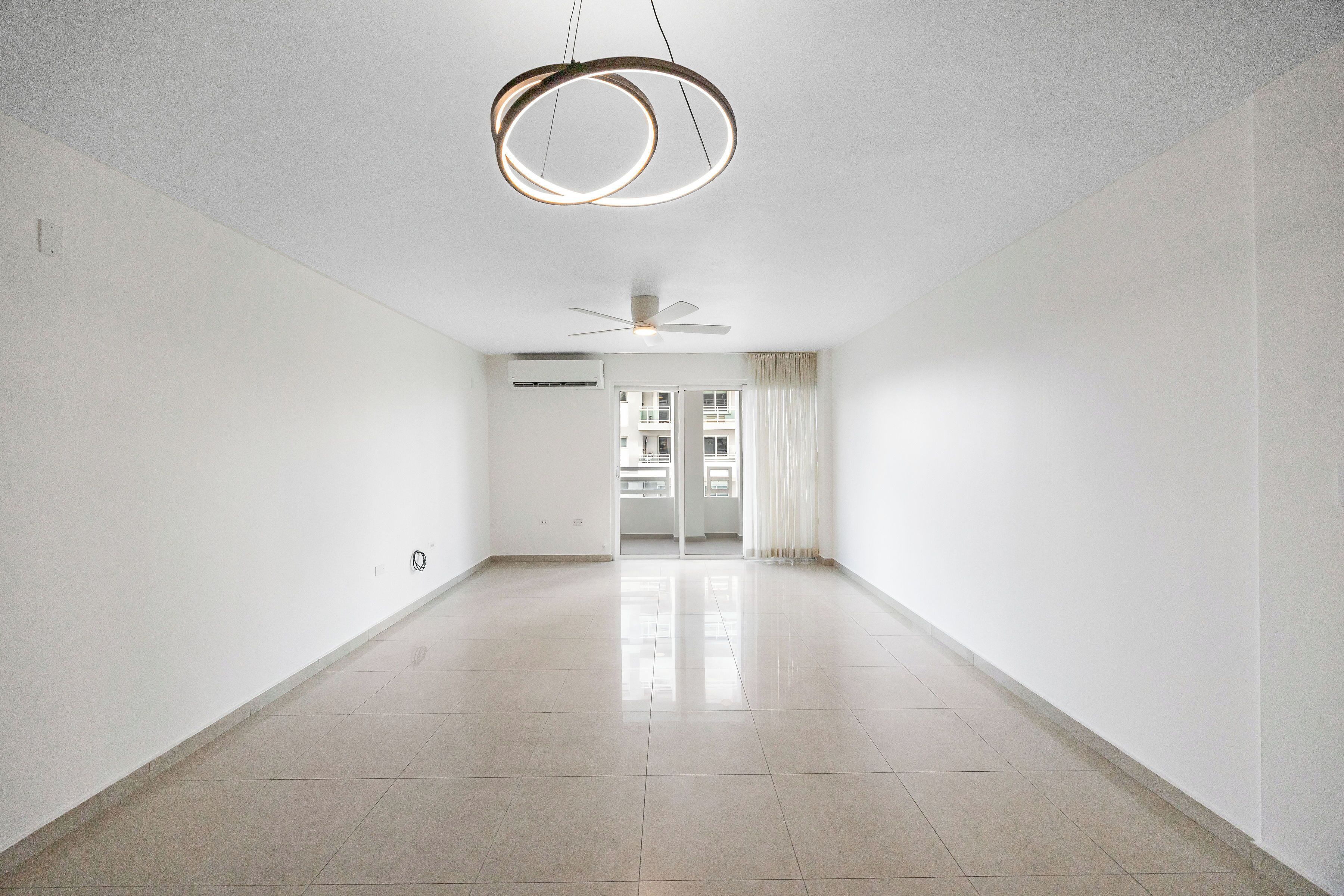 Une pièce spacieuse et bien éclairée, avec un plafond haut, un ventilateur de plafond et une vue sur une autre pièce, accessible par une porte.