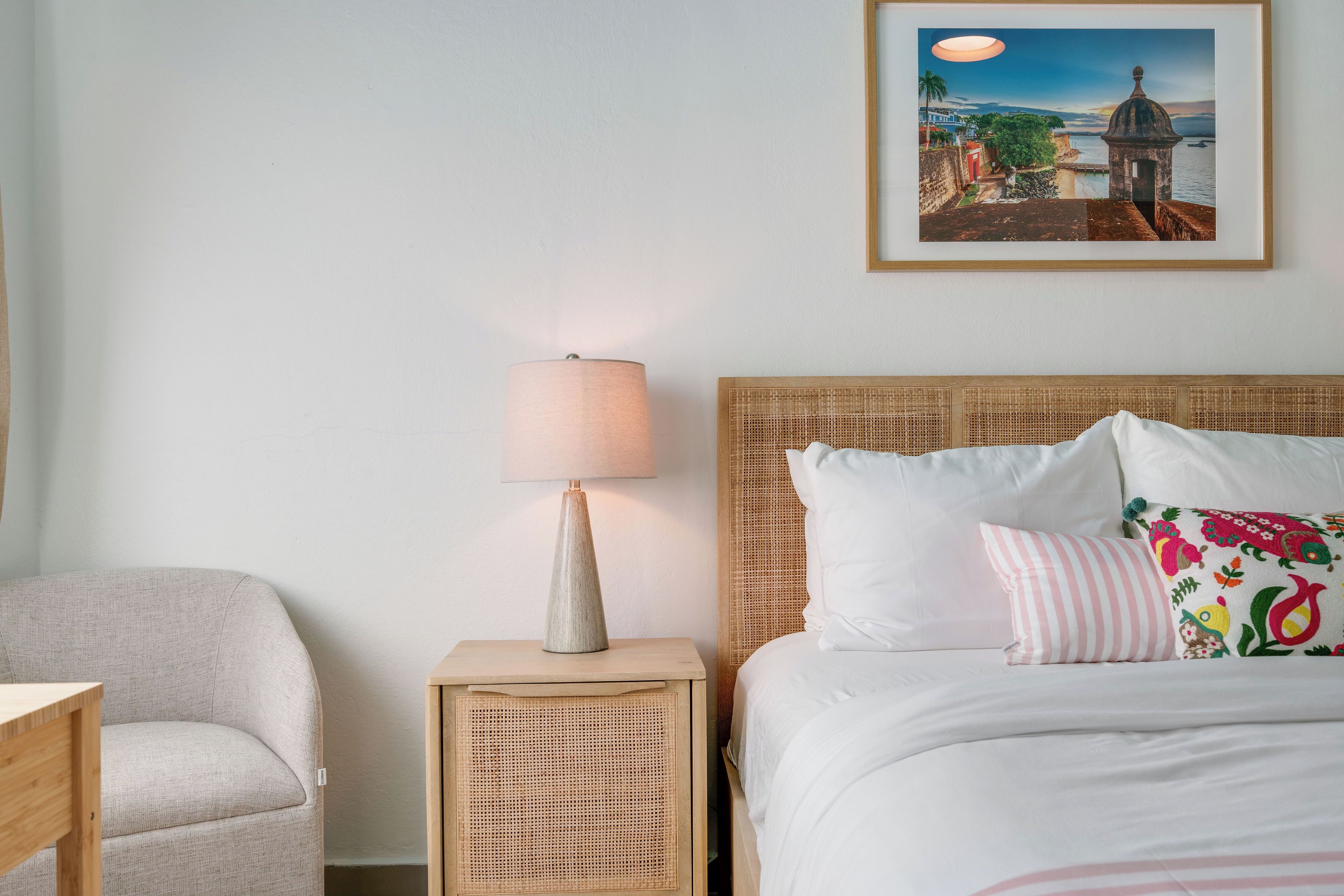 Un cuarto de hotel ordenado con cama, mesita de noche con lámpara, mesita auxiliar de mimbre, una silla y un cuadro en la pared.