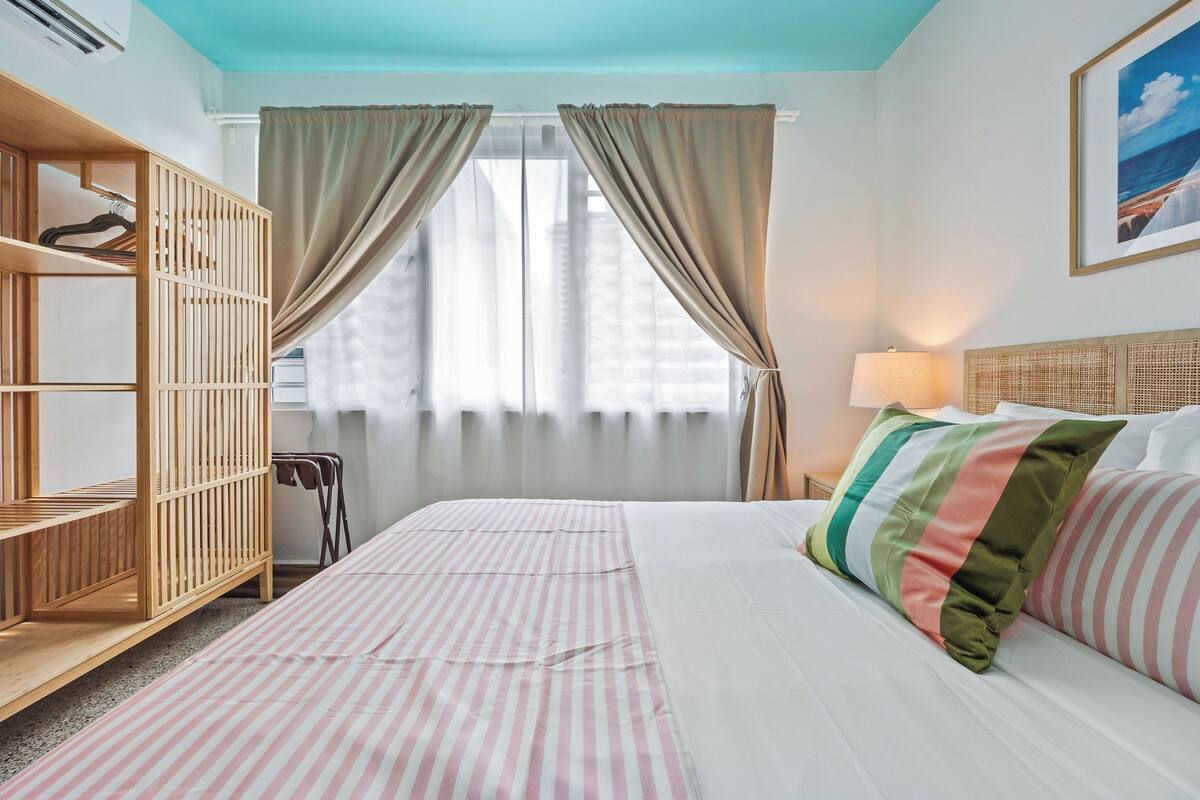Une chambre d’hôtel avec un lit, une armoire en bois, une fenêtre avec des rideaux et une lampe fixée au mur.