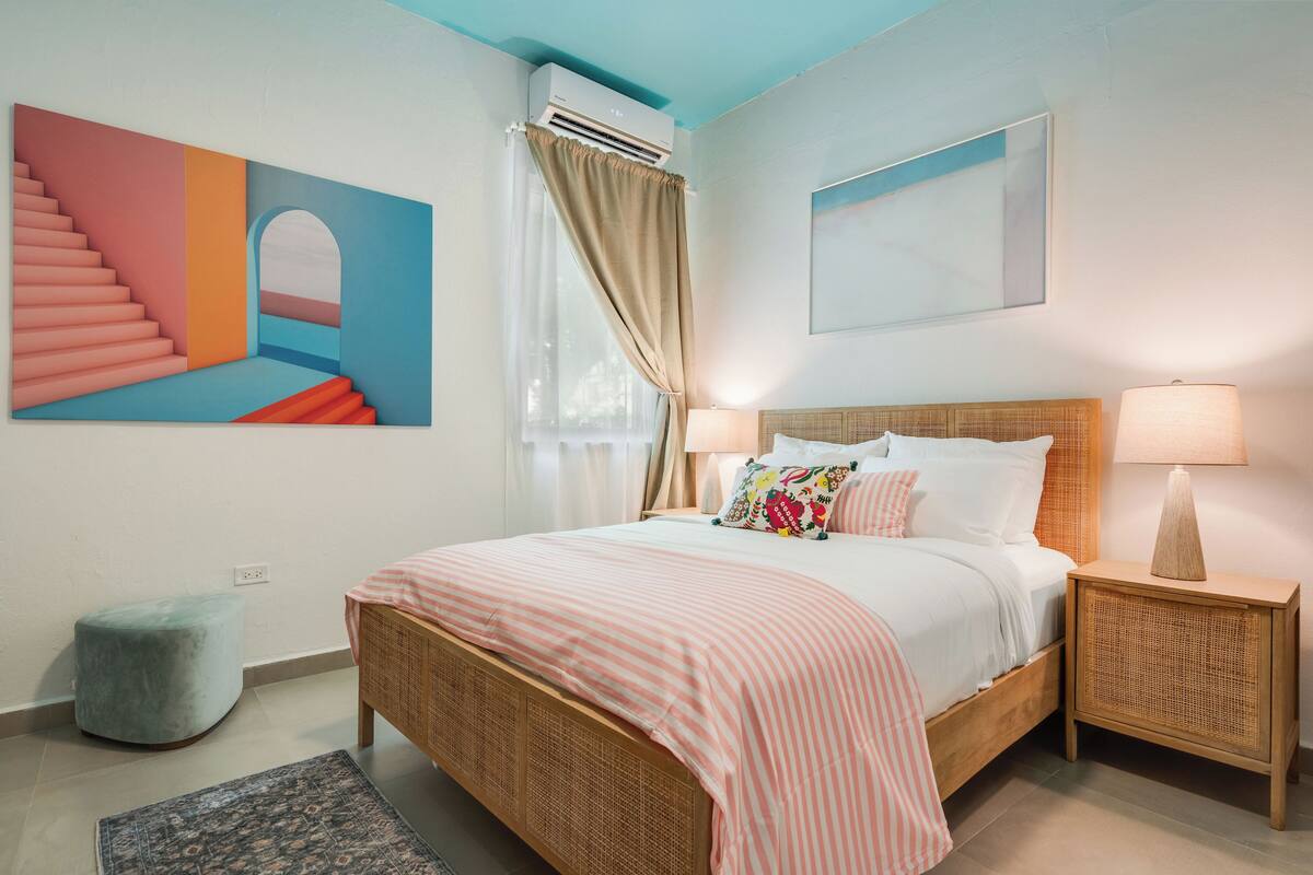 Une chambre à coucher comprenant un lit, des tables de chevet, une fenêtre avec des rideaux, un climatiseur fixé au mur et un grand tableau abstrait.