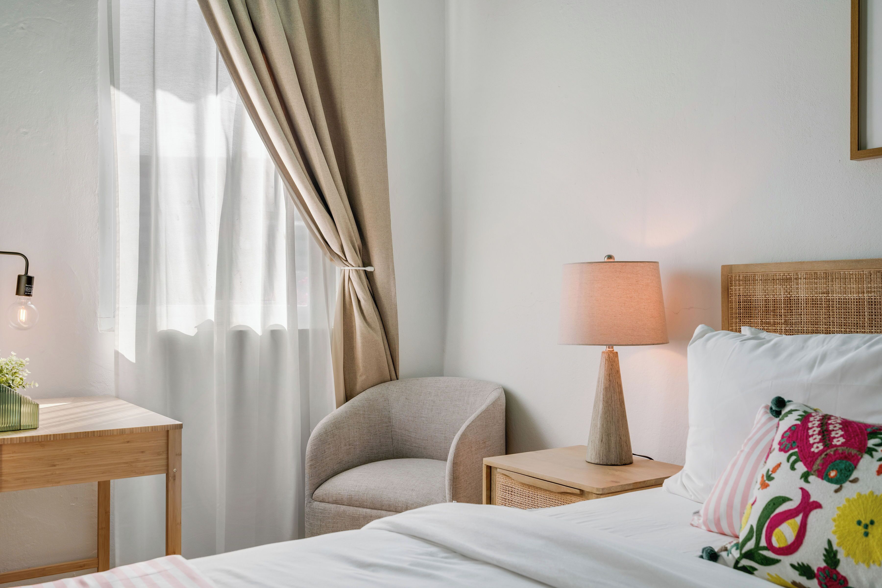 A neatly arranged hotel room with a bed, a chair, a bedside table, a lamp, and a window with curtains.