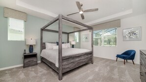 Una cama con dosel y cuatro columnas, con ventilador de techo, mesitas de noche y una silla en una habitación con ventanas amplias y obras de arte en las paredes.