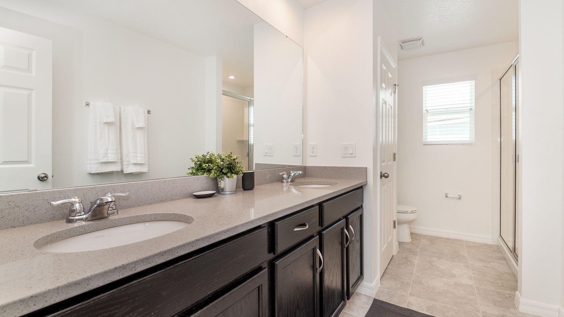 A bathroom with two sinks, a large mirror, dark cabinets, a potted plant, and a toilet.