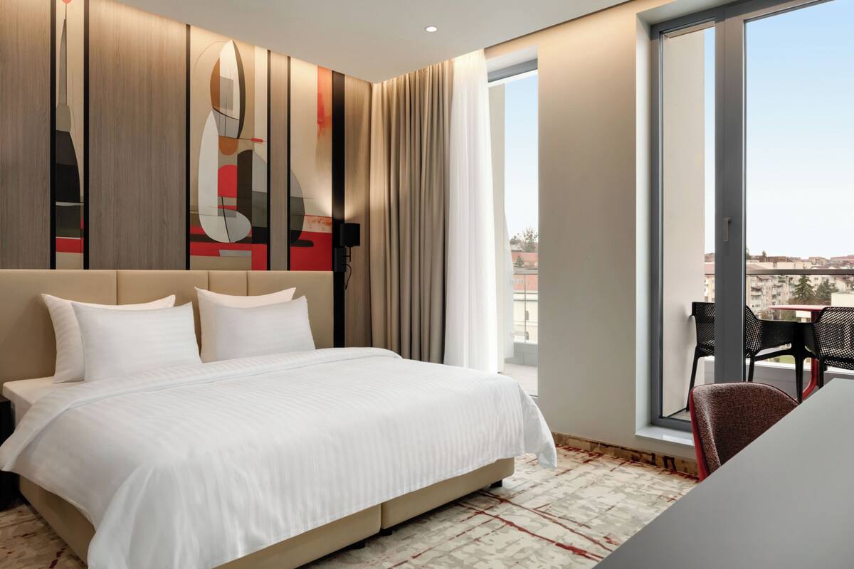 Ein Hotelzimmer mit einem großen Bett, Blick auf die Stadt und einem Balkon mit Tisch und Stühlen.