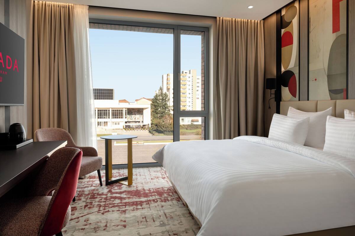 Ein Hotelzimmer mit einem großen Bett, einem Schreibtisch, einem Sessel und Blick auf Gebäude durch ein großes Fenster.
