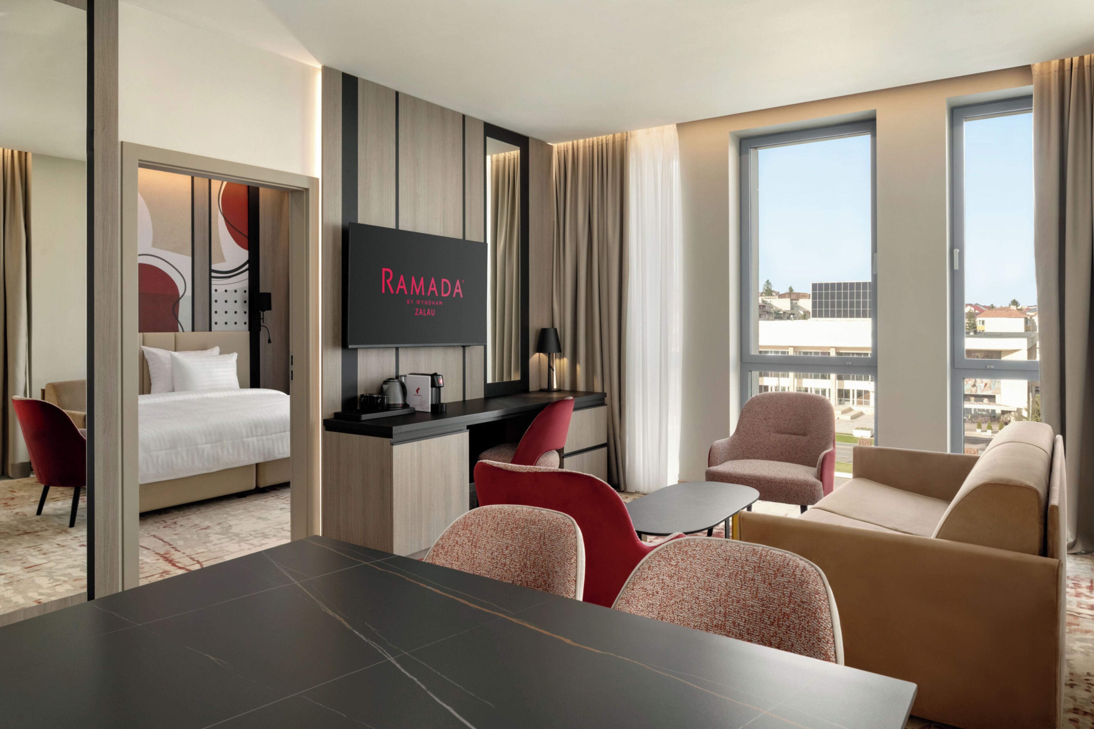 A modern hotel room with a bed, a desk with a flat-screen TV, a seating area with chairs, and a large window offering a city view.