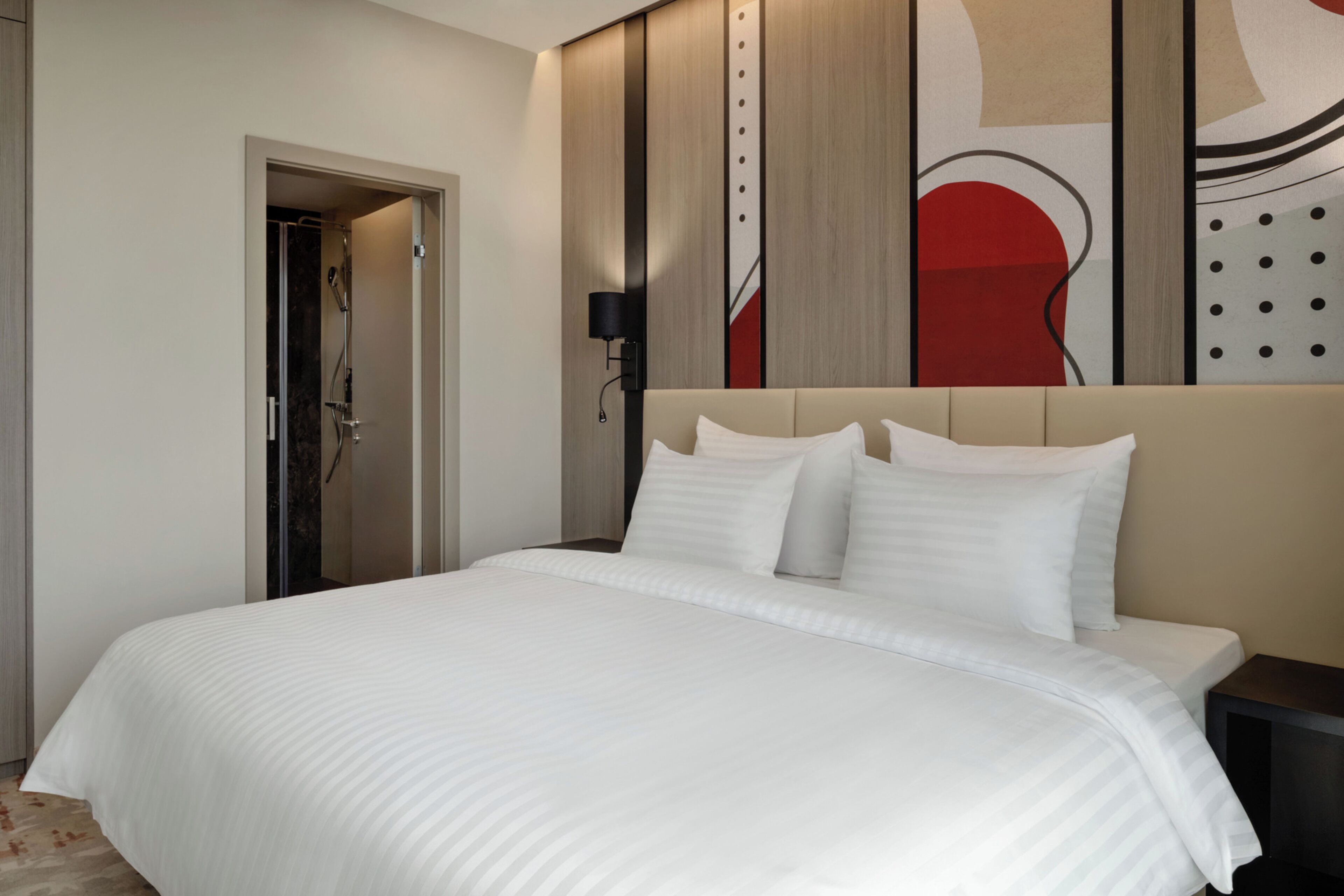 A hotel room with a bed, a shower, a wall with abstract art, and a nightstand.
