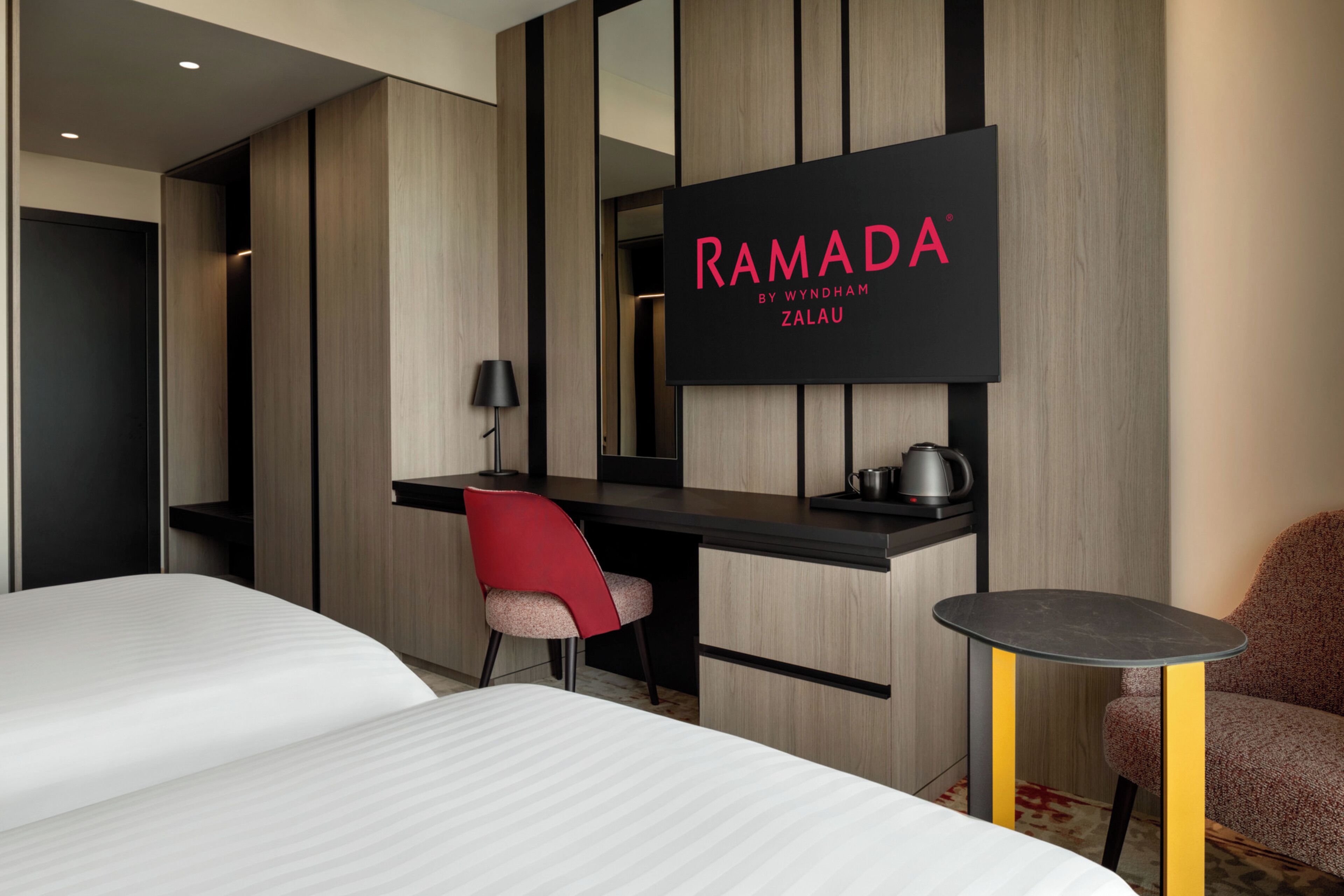 Una habitación de hotel moderna con una cama, un escritorio, una silla, una mesita y un televisor que muestra 'RAMADA BY WYNDHAM ZALAU'.