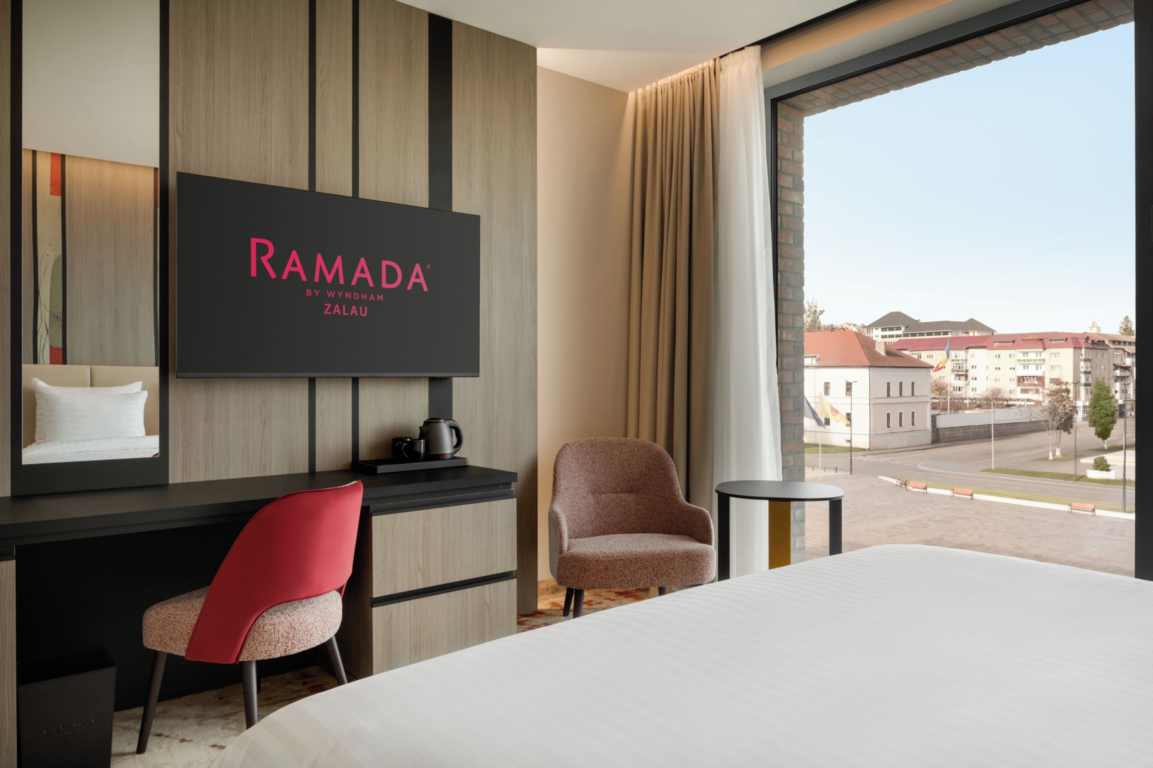 Una habitación de hotel con cama, escritorio, silla, televisor mostrando 'RAMADA BY WYNDHAM ZALAU' y vistas a la ciudad a través de amplios ventanales.