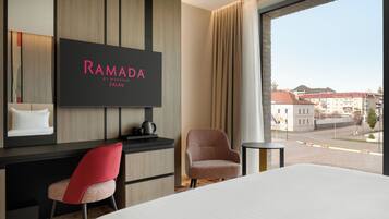 Una habitación de hotel con cama, escritorio, silla, televisor mostrando 'RAMADA BY WYNDHAM ZALAU' y vistas a la ciudad a través de amplios ventanales.