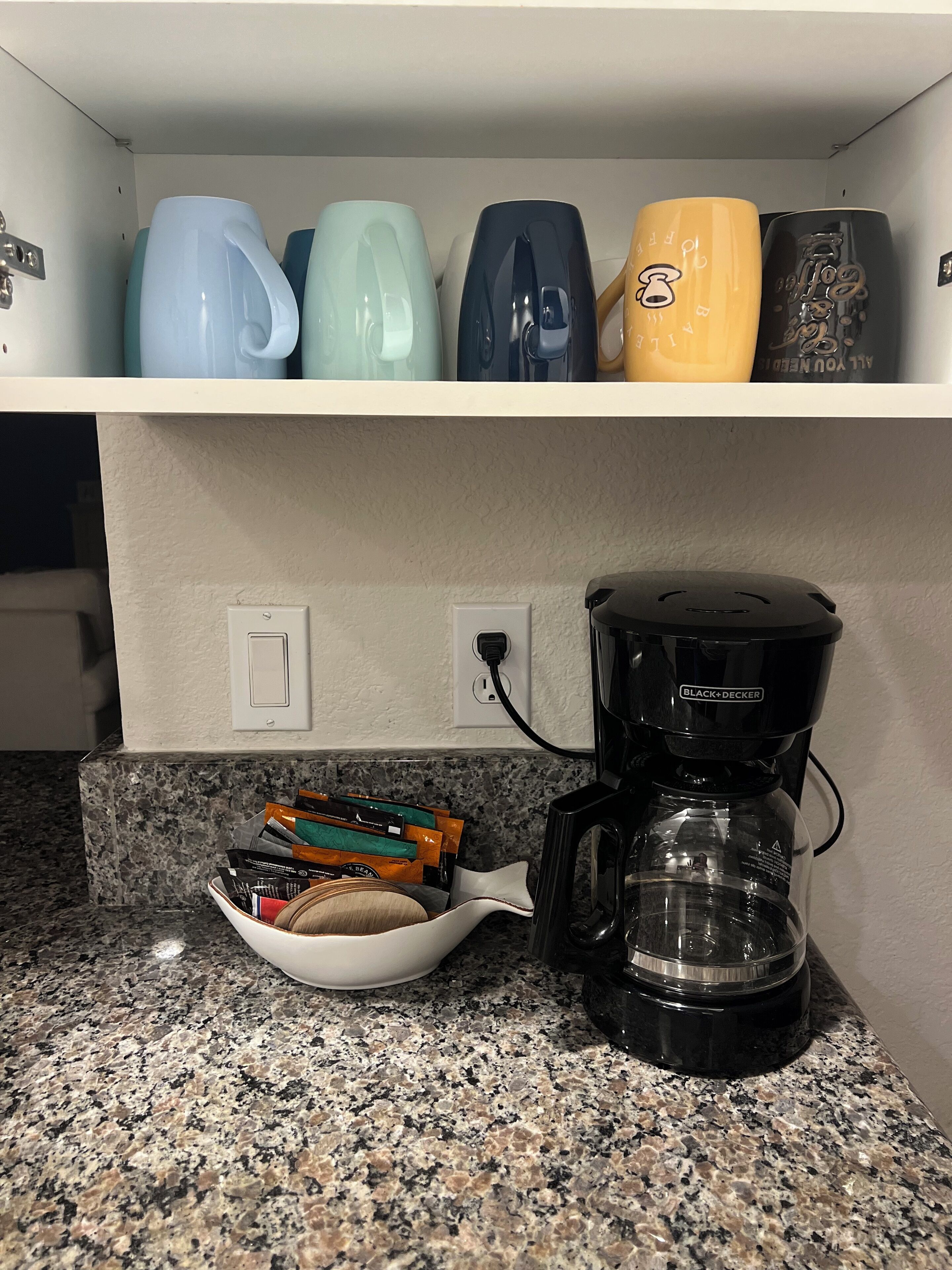 Een aanrecht met een koffiezetapparaat, een kom koffiecups en een plank met verschillende mokken.