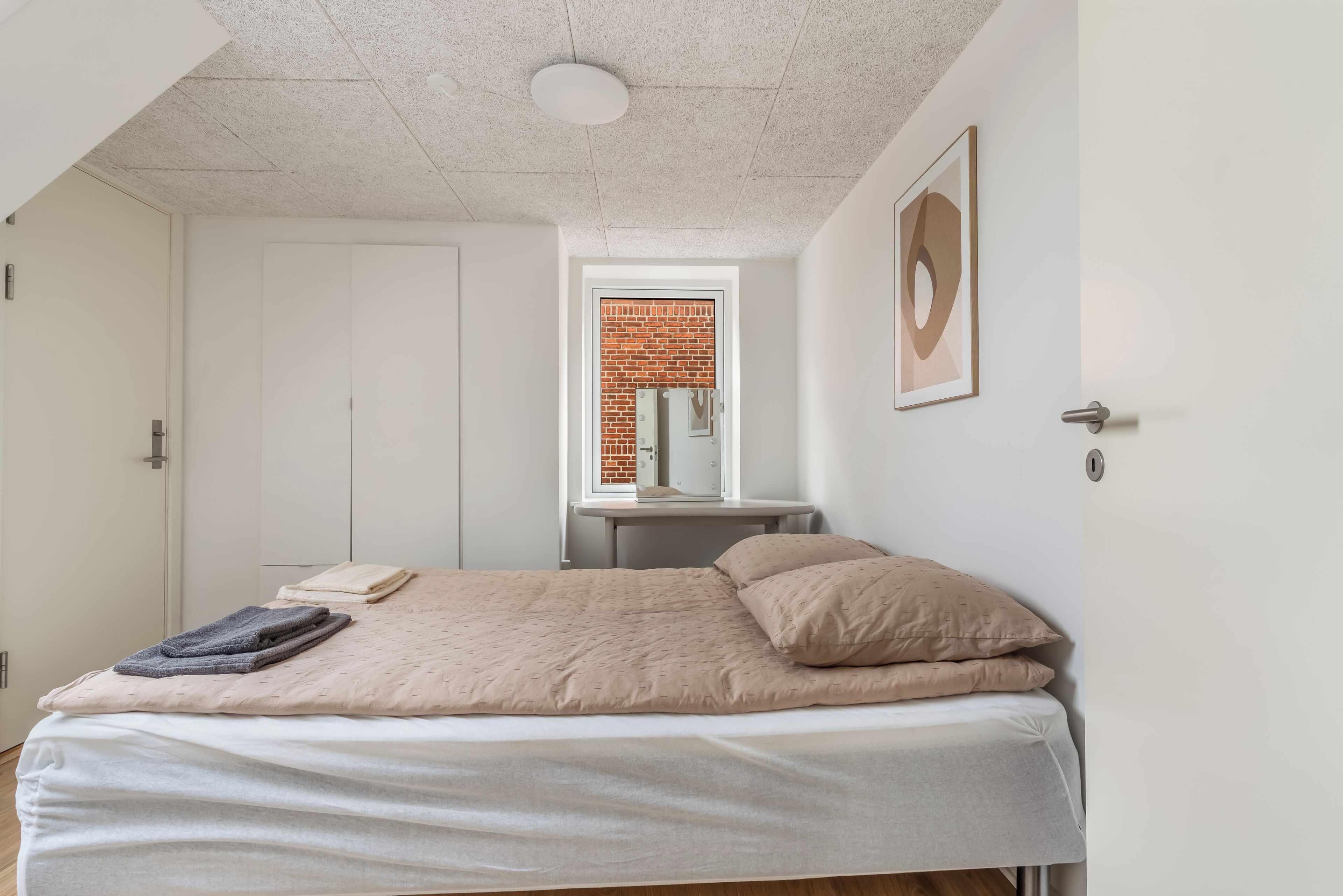 A bedroom with a bed, a window with a view of a brick wall, a door, and a framed picture on the wall.