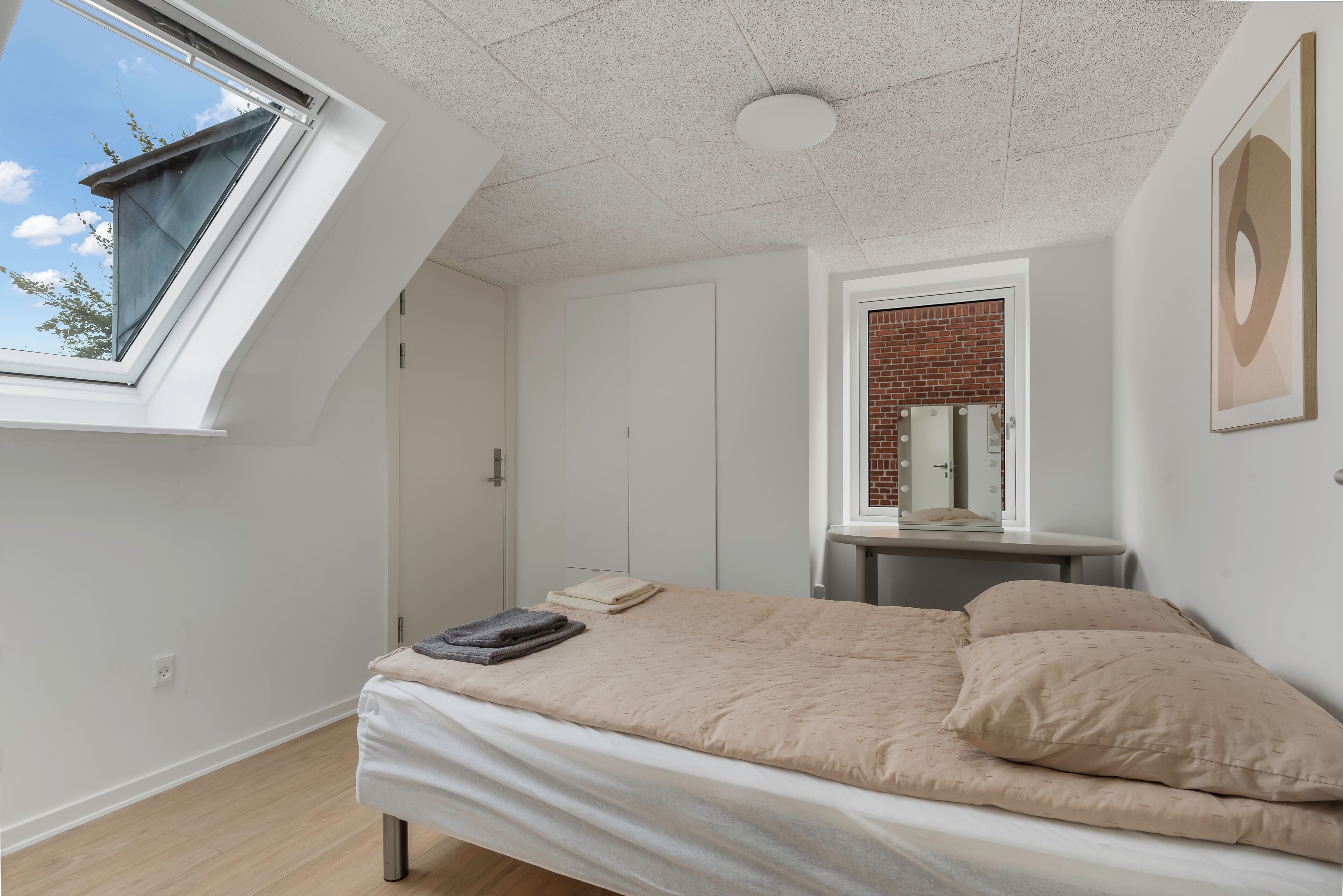 A bedroom with a skylight, a bed with beige bedding, a small table, and a mirror with a brick wall backdrop.