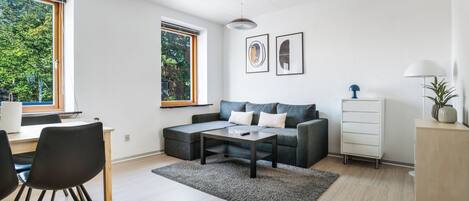A modern living room with a sofa, coffee table, and a desk with chairs. There are two windows with views of trees.