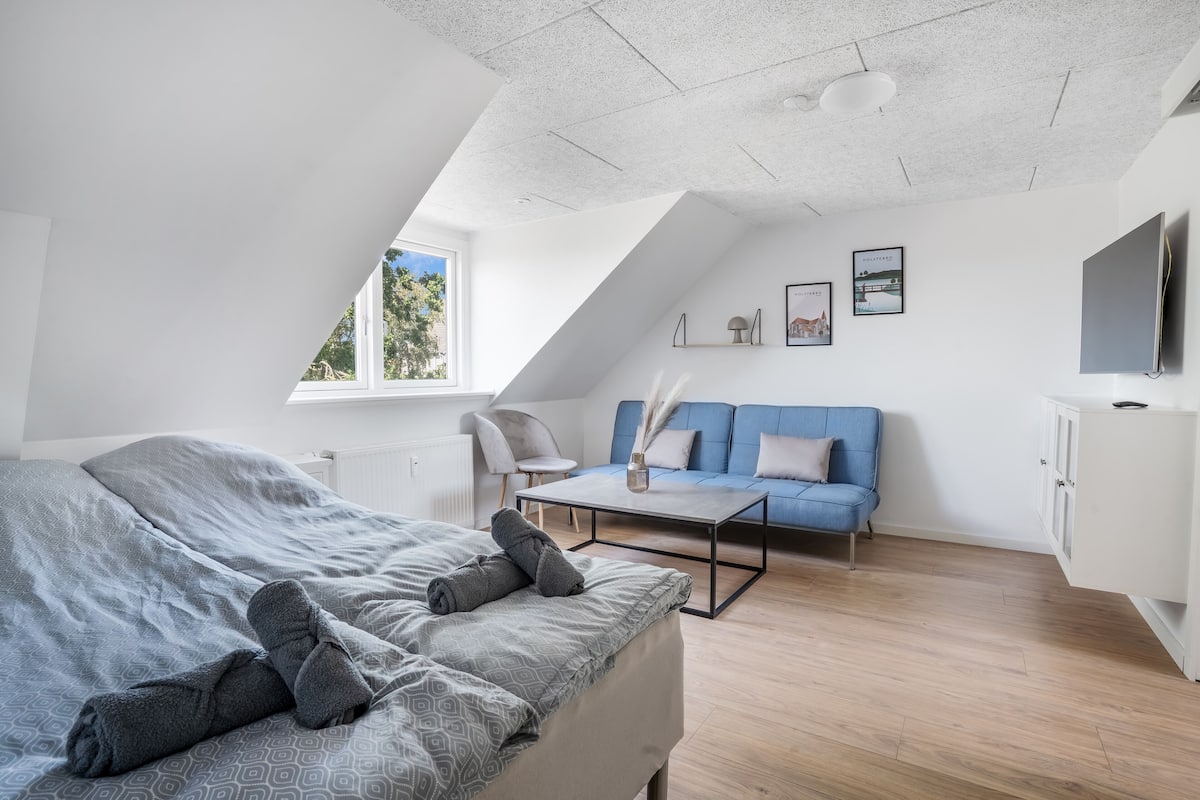 A modern loft bedroom with a sloped ceiling, a bed with grey bedding, a blue sofa, a small table, and a TV on the wall.