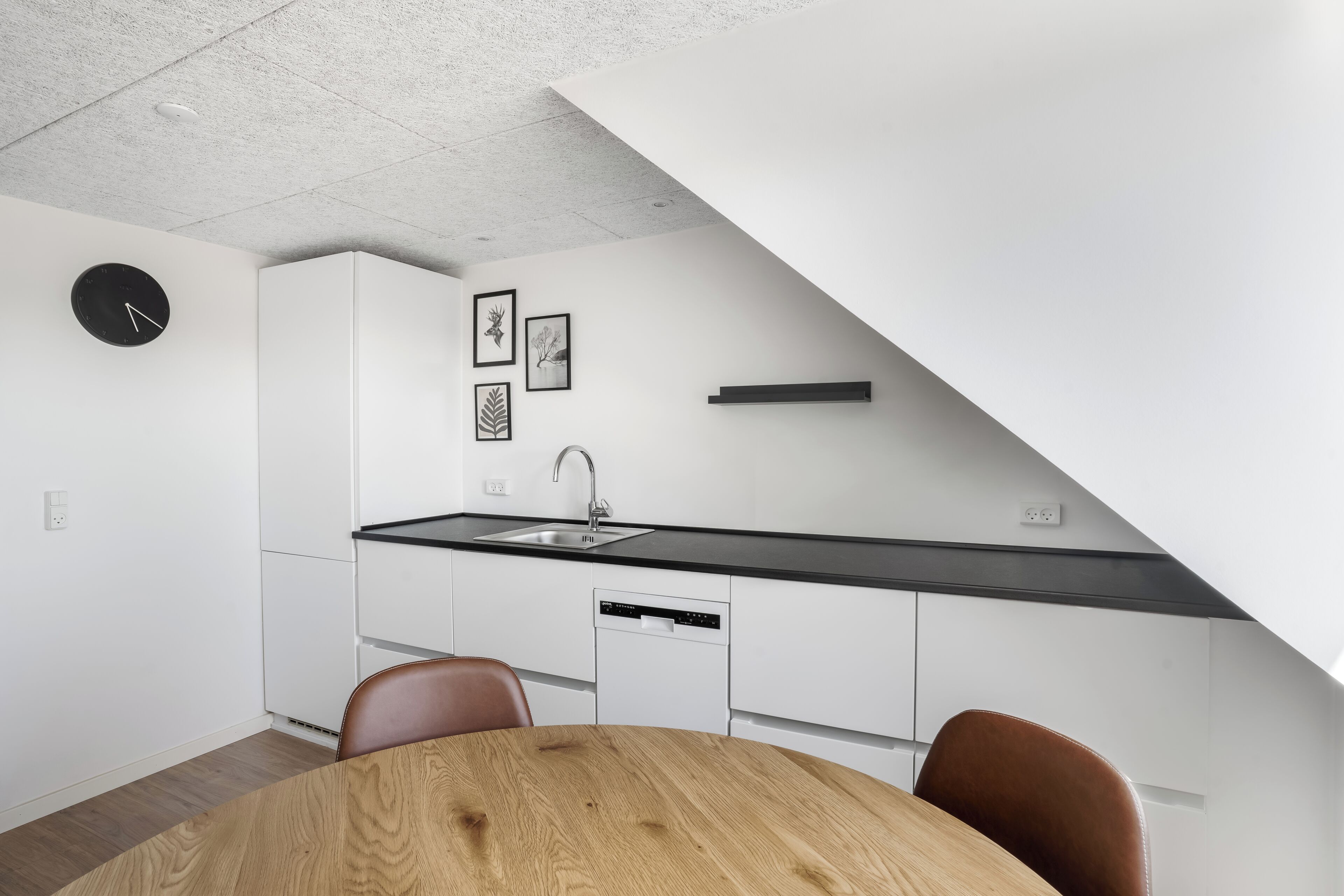 Une cuisine moderne avec des meubles blancs, un plan de travail noir, une horloge et des reproductions botaniques encadrées.