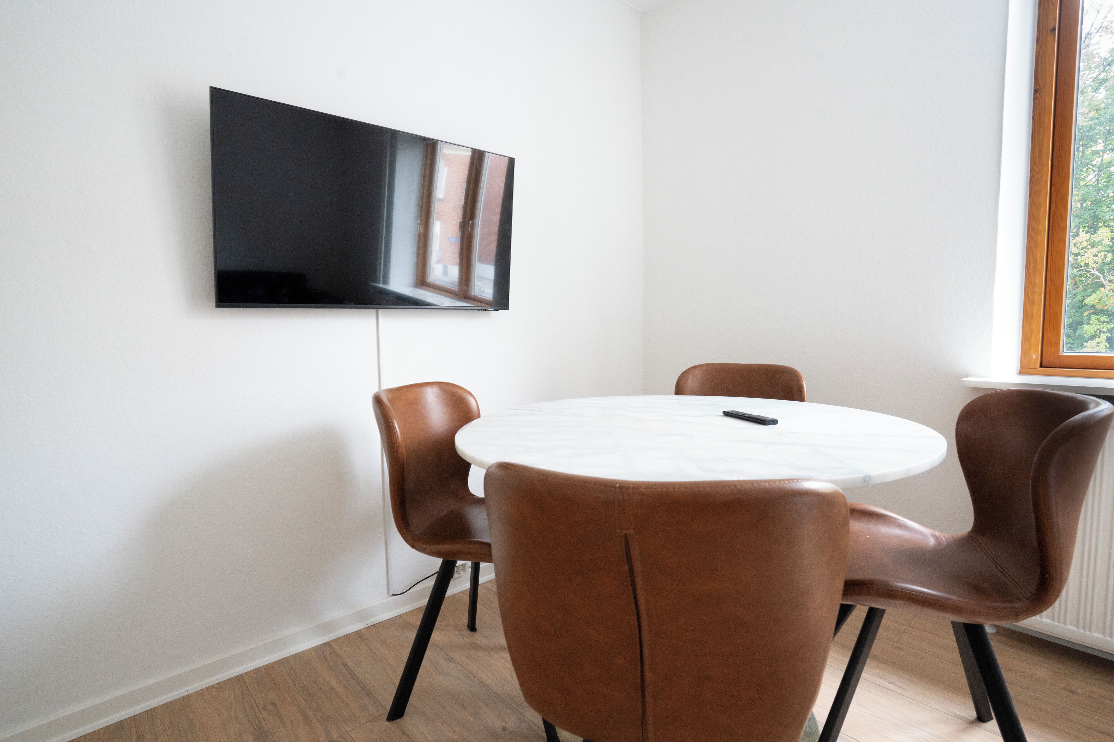 A modern dining area with a round table and four brown chairs, a mounted flat-screen TV on the wall, and a window with a view of trees.