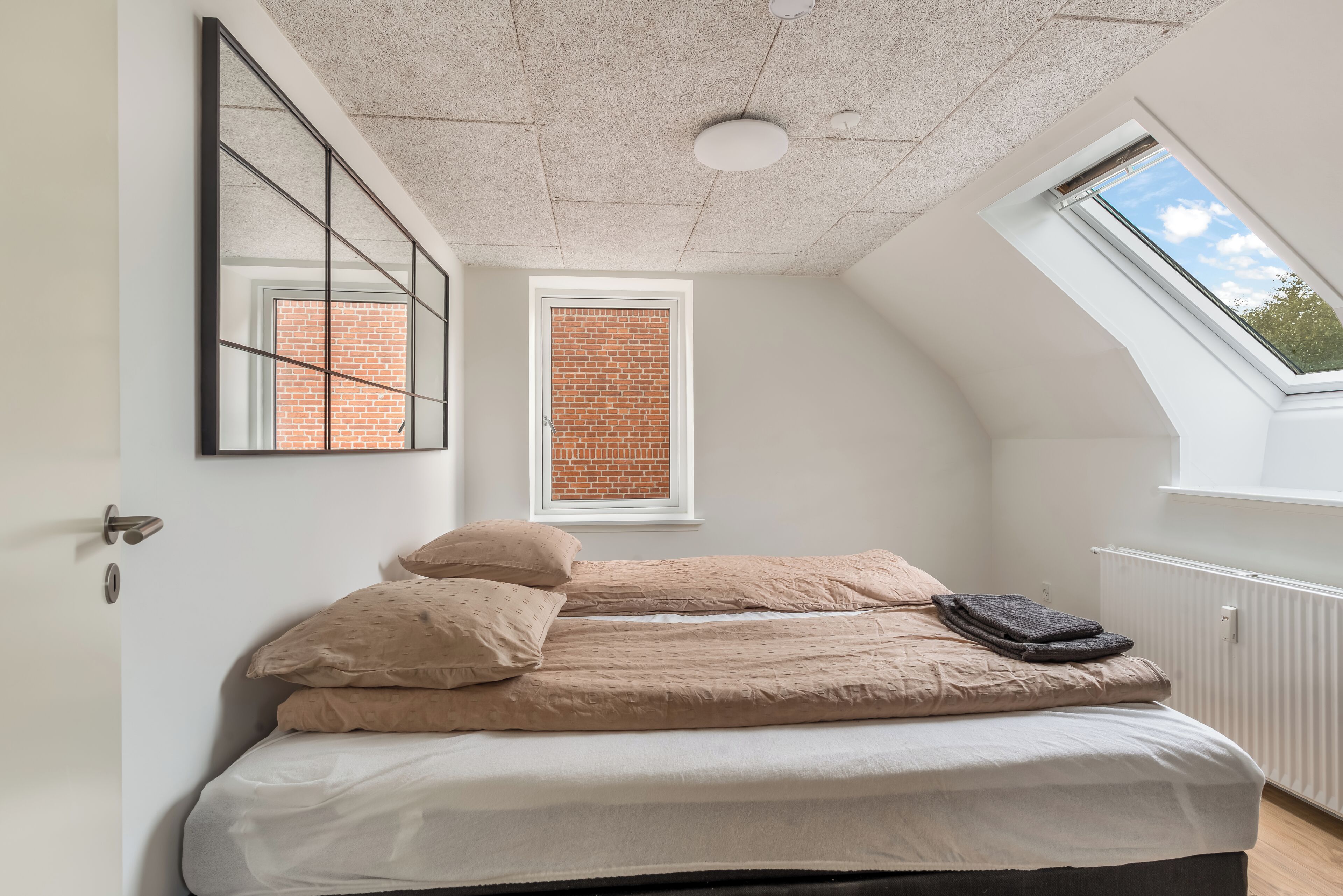Ein Schlafzimmer mit schrägverdeckter Decke, einem Dachfenster, einem Bett mit beigefarbener Bettwäsche, einem Fenster mit Blick auf eine Ziegelwand und einem Heizkörper.