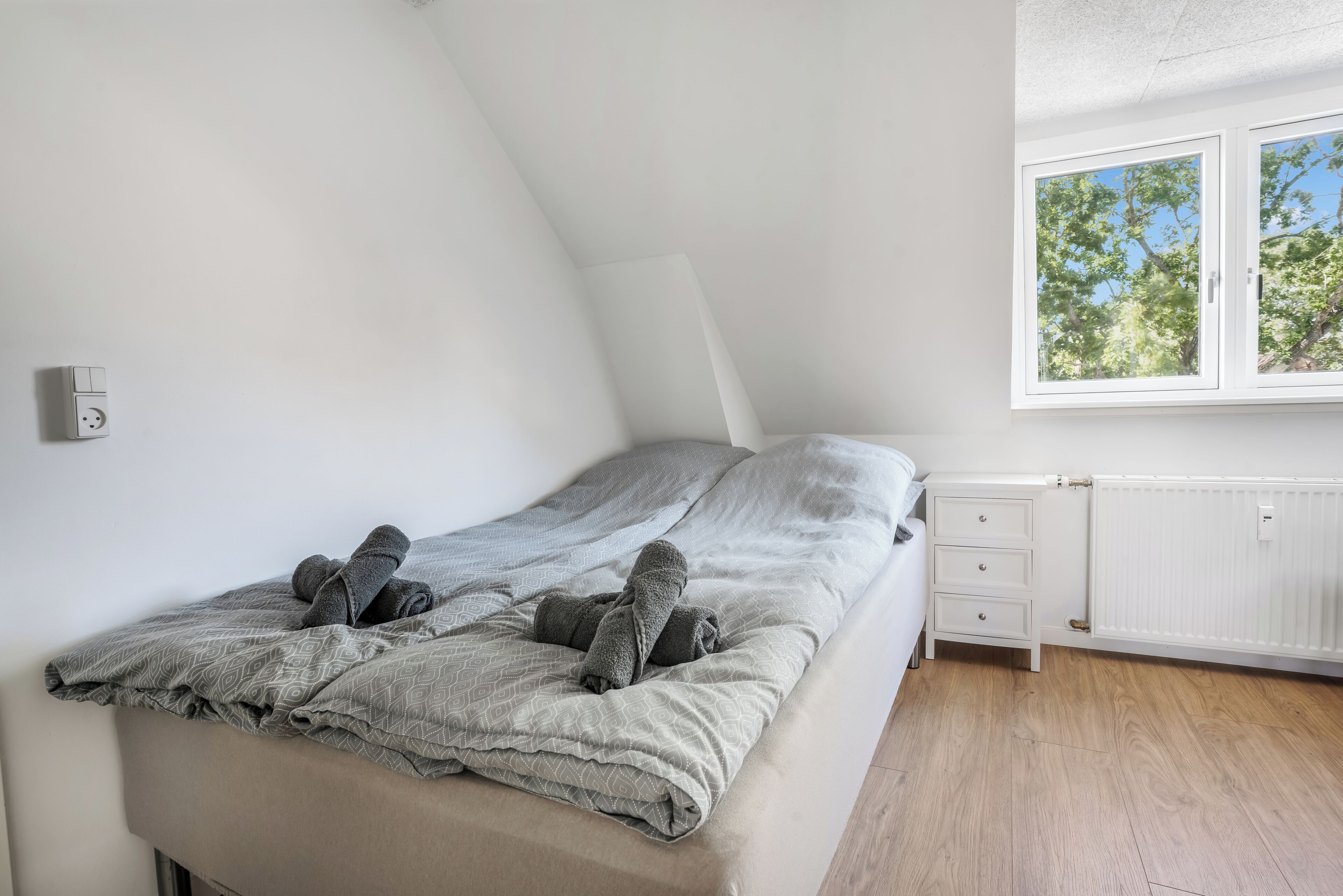 Un dormitorio con techo inclinado, una cama con ropa de cama gris, un tocador blanco y una ventana con vista a árboles.