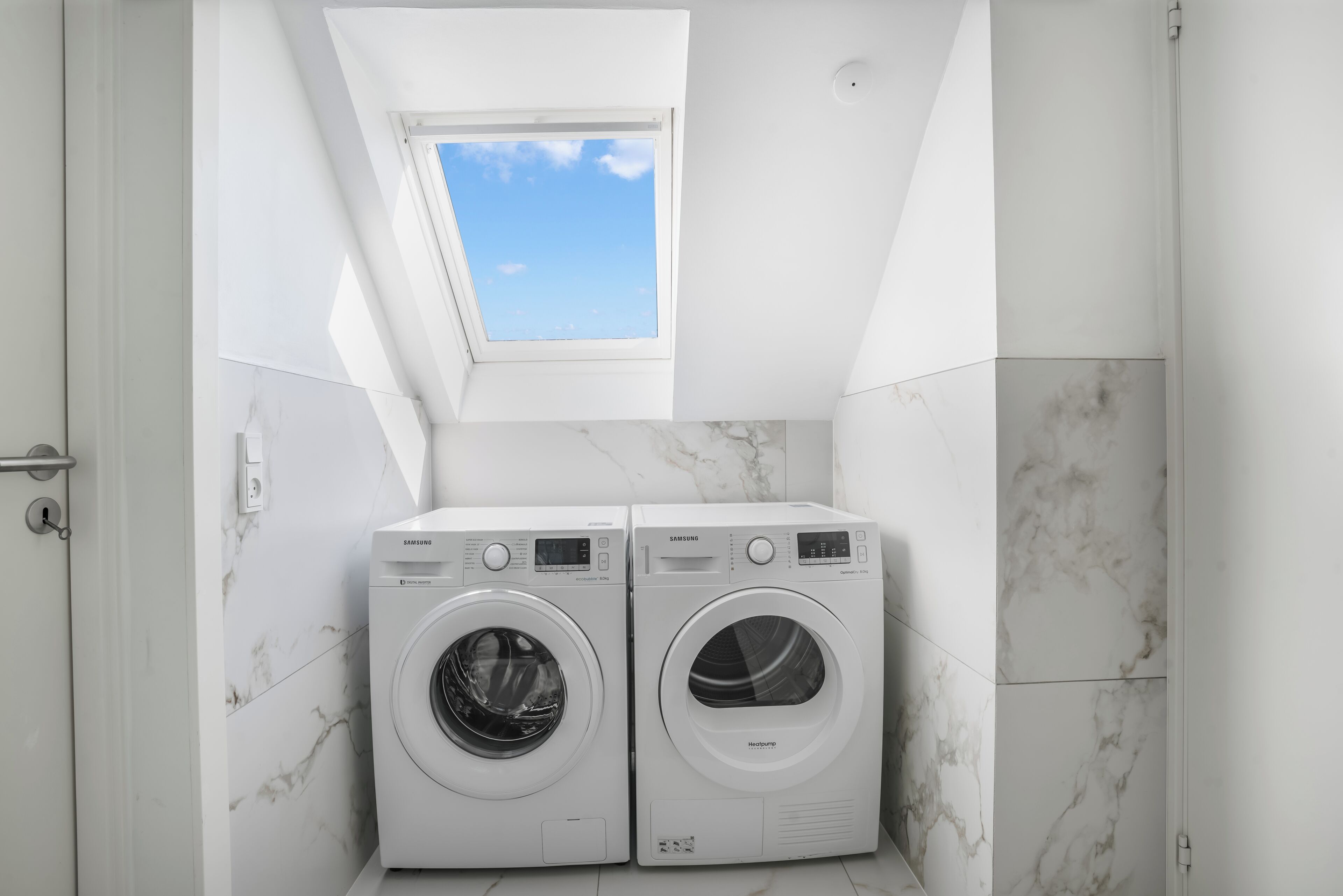 Un lavadero con dos lavadoras Samsung, un tragaluz y paredes con azulejos de mármol.