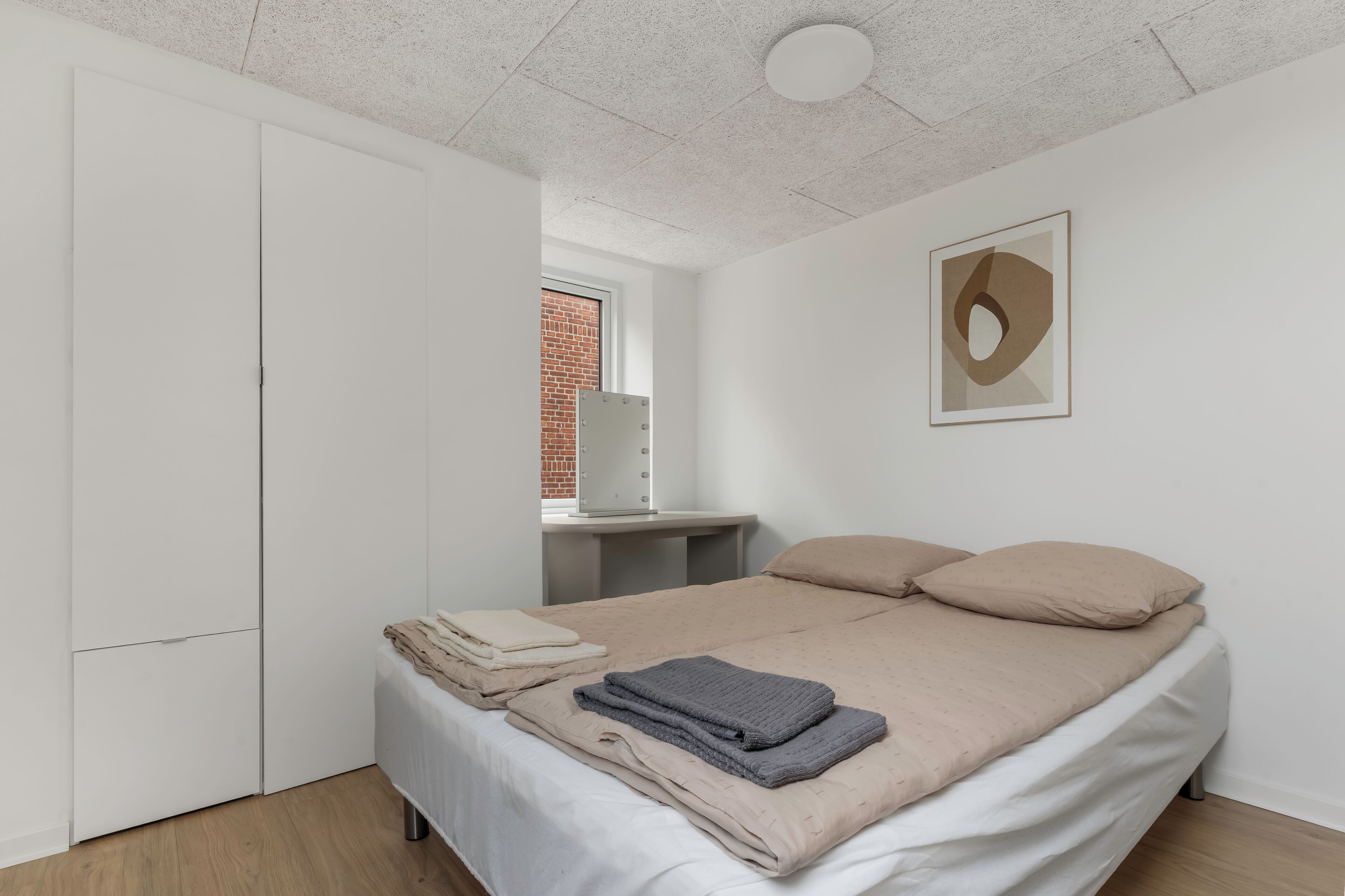 A bedroom with a bed, a wardrobe, a vanity with a mirror, and a framed abstract artwork on the wall.