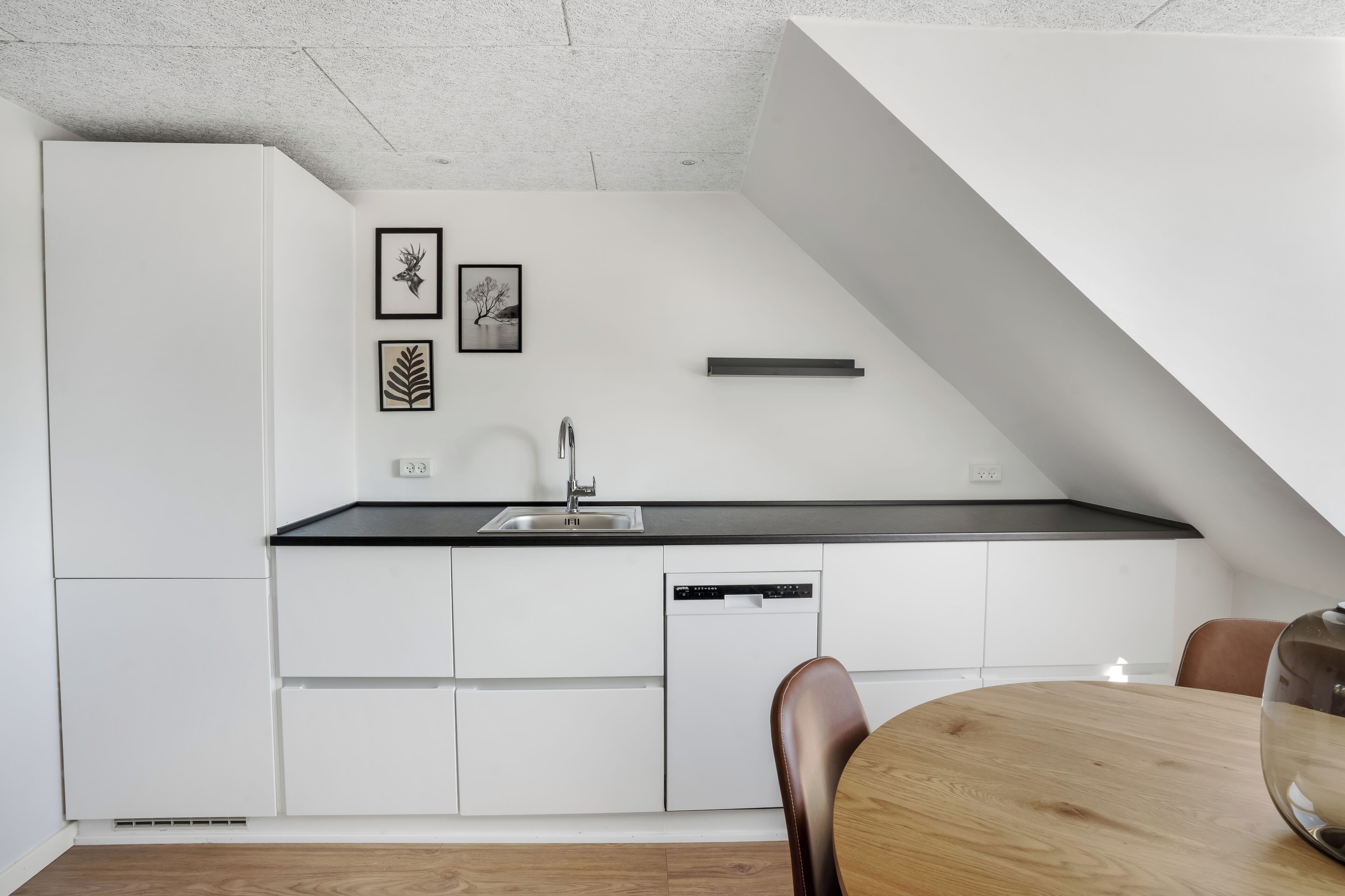 A modern kitchen with white cabinetry, a black countertop, a dishwasher, and a dining area with a wooden table and chairs.