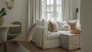 Un salón acogedor con un sofá blanco, una mesita auxiliar, sillas de mimbre y vistas a un edificio a través de amplios ventanales.