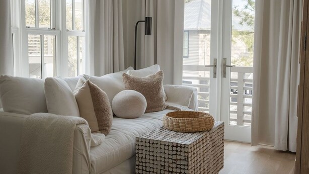 Un sofá blanco con cojines, una cesta tejida y una mesita lateral estampada en una habitación luminosa con grandes ventanales y vistas a un edificio.