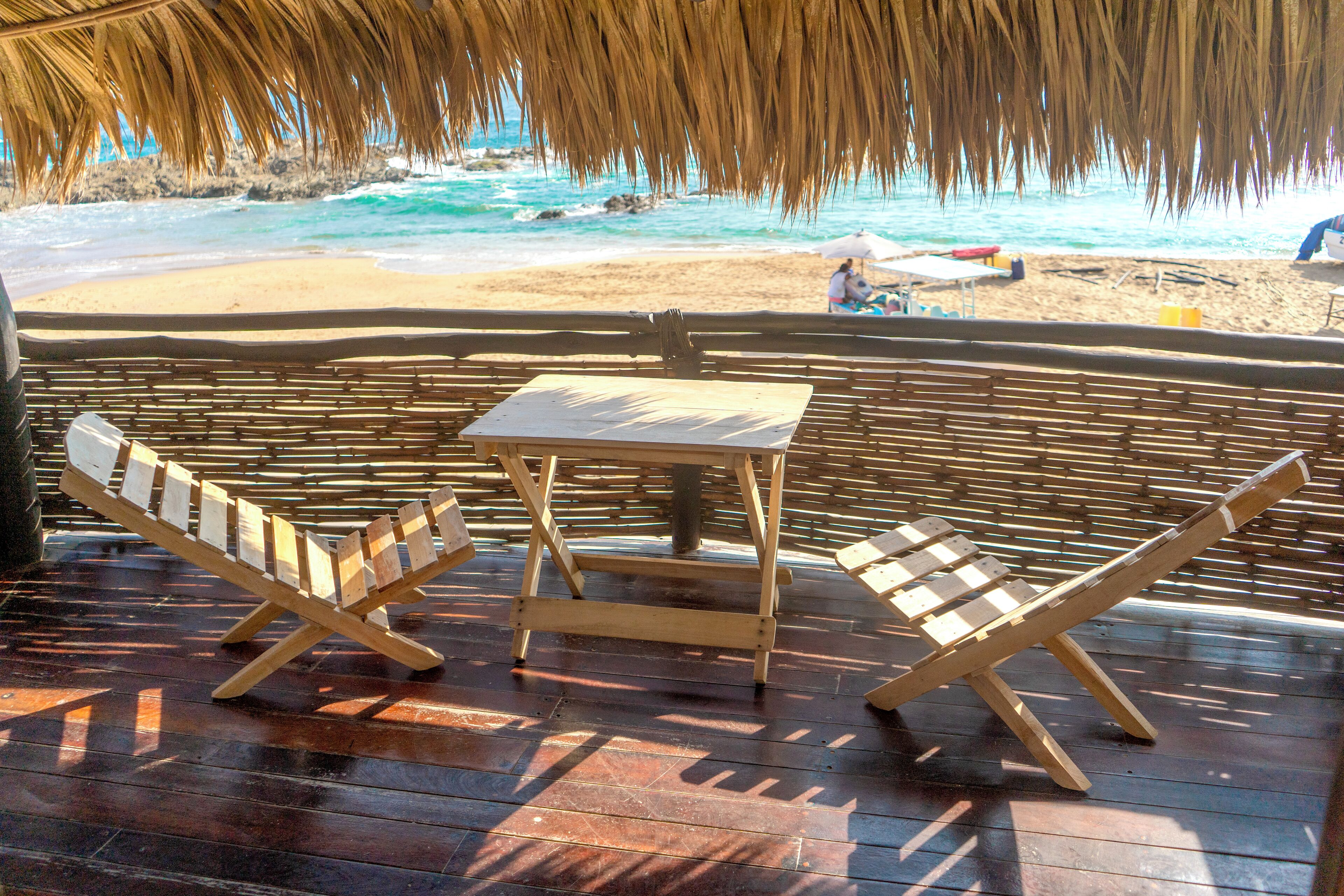Terraza de madera con dos sillas plegables y una pequeña mesa, con vista a una playa y una estructura con techo de paja.