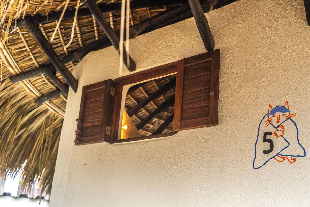 Una habitación con techo de paja rústico, una ventana de madera con contraventanas, y una pared con el número 5 pintado en azul y rojo.