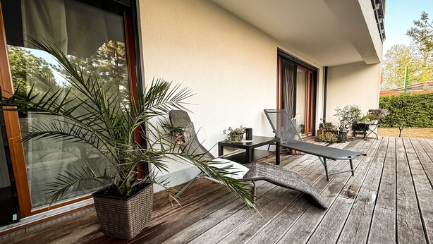 Een houten terras met buitenmeubilair, waaronder een lounge stoel en een bijzettafeltje, nabij een gebouw met grote ramen en een potplant.'