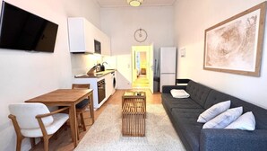 Een ruimtelijk ingerichte leefruimte met kitchenette, een eethoek, een bank en een aan de muur bevestigde televisie.
