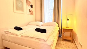 Een netjes opgemaakt bed met kussens en dekbed, een klein nachtkastje met een lamp, en kunst aan de muur.