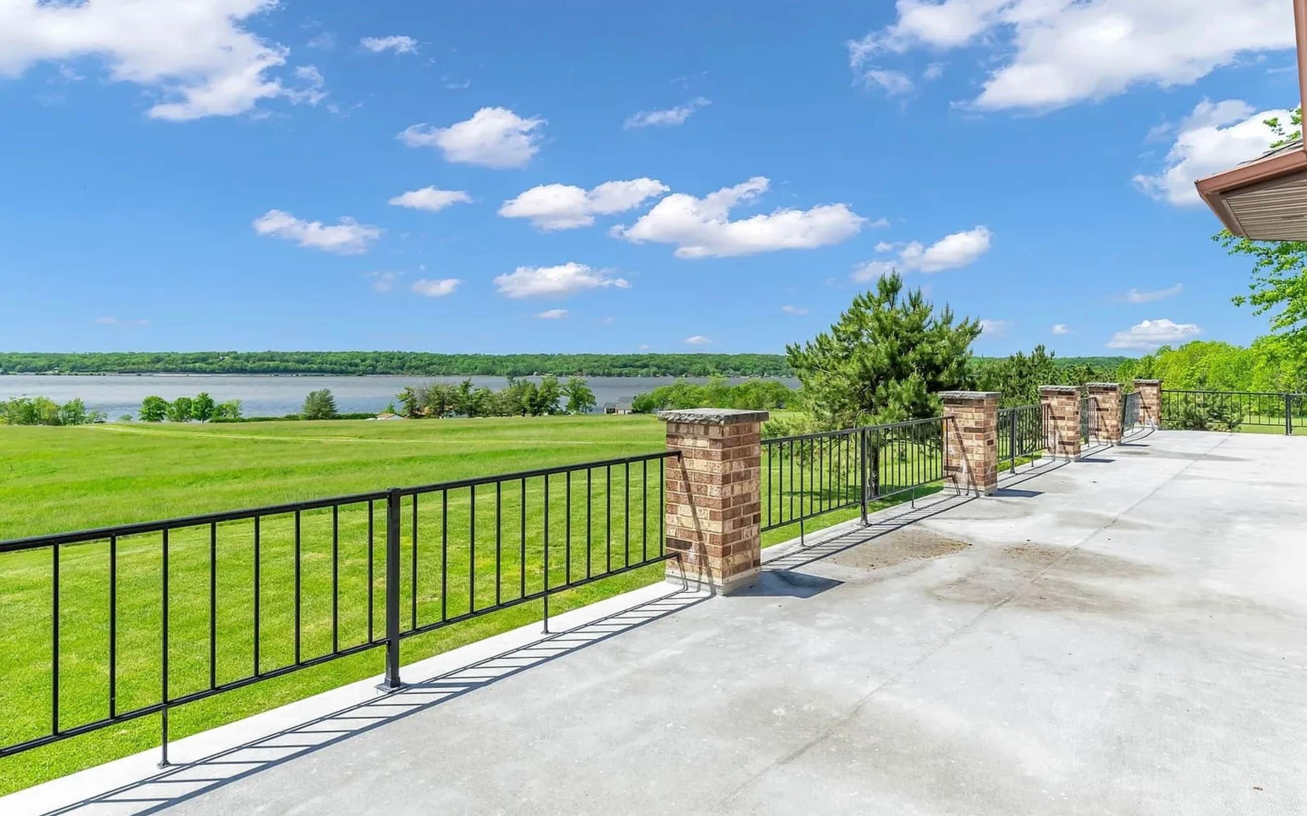 Un balcon doté d’une balustrade en métal, d’un pilier en briques et offrant une vue sur un lac et un paysage verdoyant.