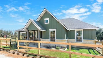 Une maison à deux étages, avec une façade verte et un toit gris, entourée d’une clôture en bois et d’un petit jardin.