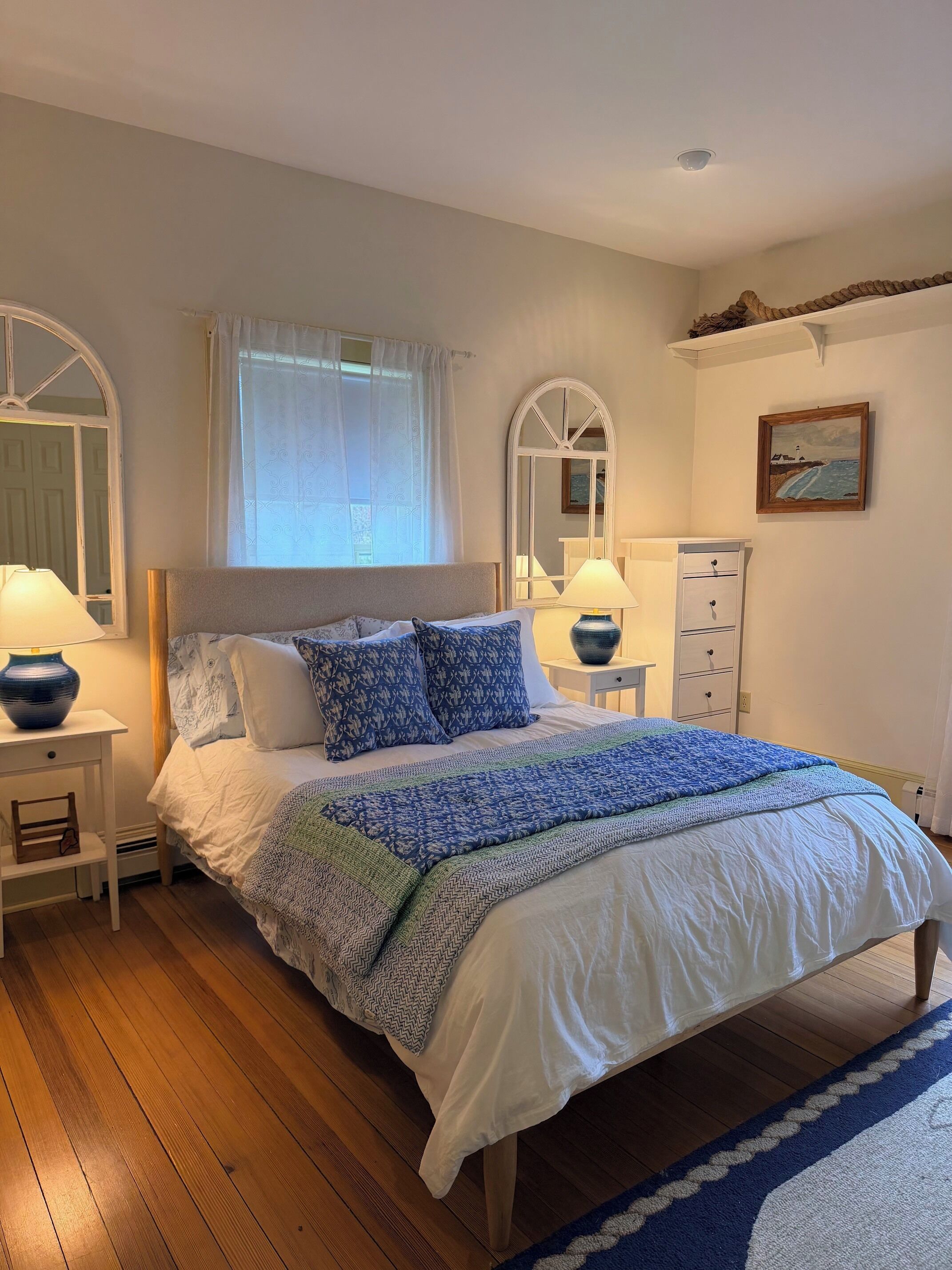 A bedroom with a bed, two bedside tables, a dresser, a window with curtains, and a painting on the wall.