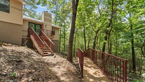A wooden staircase leading to a beige building with green doors, surrounded by a forested area.