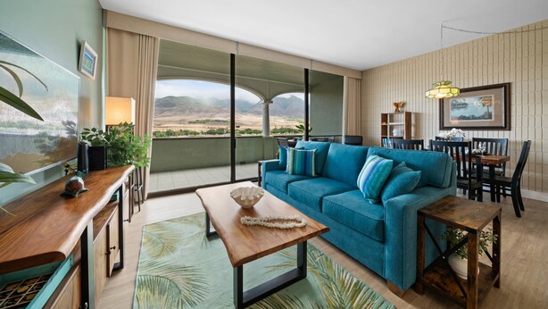 Un salón moderno con un sofá azul, una mesa de centro de madera y vistas a un paisaje montañoso a través de amplios ventanales.