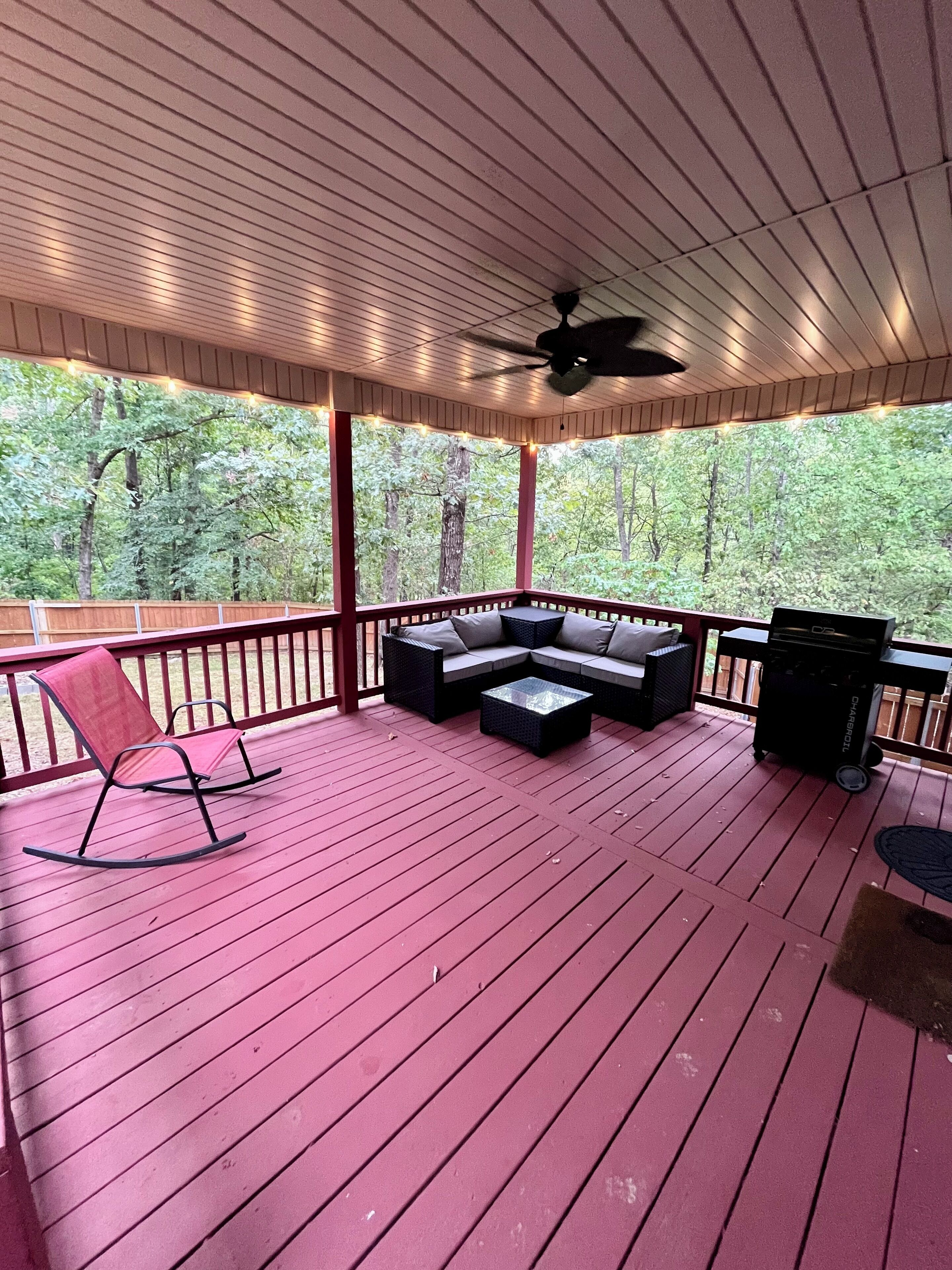 A covered deck with a red floor, a ceiling fan, string lights, a sofa, a rocking chair, and a grill.
