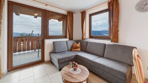 A living room with a grey sofa, a small round table with a vase, and large windows with curtains. - Apartment 'Adlerhorst' with Mountain View, Shared Garden and Wi-Fi (Lagundo)