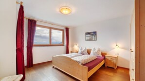 A bedroom with a wooden bed, a nightstand, a window with curtains, a wall-mounted light, and a small table. - Apartment 'Schwalbennest' with Mountain View, Shared Garden and Wi-Fi (Lagundo)