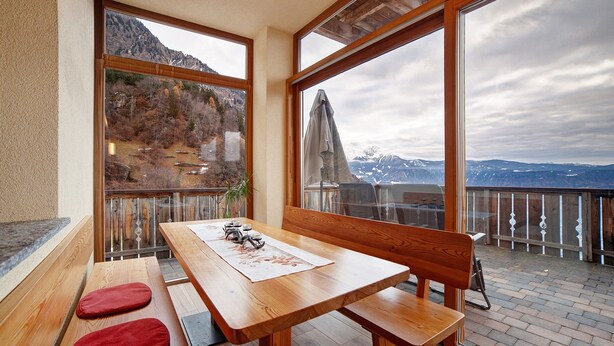 A wooden table with benches, a view of mountains through large windows, and a balcony with a railing. - Apartment 'Storchennest' with Mountain View, Shared Garden and Wi-Fi (Lagundo)