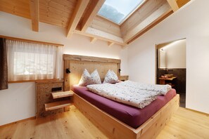 Een houten bed met schuine daklijn en dakraam, een houten nachtkastje en een raam met dunne gordijnen.