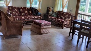 Una habitación amplia con techo alto y vigas a la vista, amueblada con un conjunto de sofás y sillas de mimbre, una mesa de comedor y un ventilador de techo.