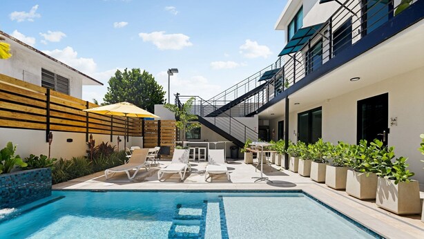 A swimming pool with lounge chairs and umbrellas, surrounded by potted plants and a modern building with balconies. - Isla Verde Ocean View | Entire 2nd Floor Suite (San Juan)