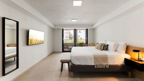 A modern bedroom with a large bed, a bench, a bedside table with a lamp, a TV, and a view of the outdoors through a glass door. - Isla Verde Ocean View | Entire 2nd Floor Suite (San Juan)