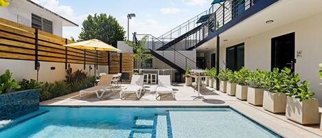 A swimming pool with lounge chairs and umbrellas, surrounded by potted plants and a modern building with balconies.